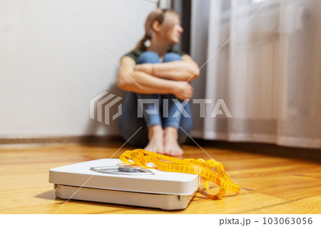 White scale with measuring tape and depression, upset woman sitting on floor White scale with measuring tape and depression, upset woman sitting on floor 103063056