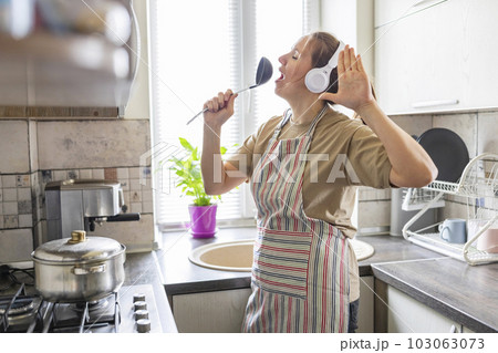 Woman in earphones listening to music and singing while cooking in the kitchen 103063073