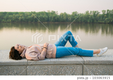 Woman lying down on embankment by lake or river at city park - dreamer and relax concept Woman lying down on embankment by lake or river at city park - dreamer and relax concept 103063915