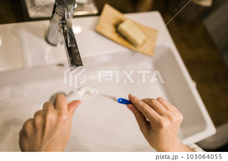 Female hand holding toothbrush with toothpaste applied on it in bathroom. Close up of female hand ready for brushing teeth. Young woman hand holding toothbrush with white tooth paste. Female hand holding toothbrush with toothpaste applied on it in bathroom. Close up of female hand ready for brushing teeth. Young woman hand holding toothbrush with white tooth paste. 103064055