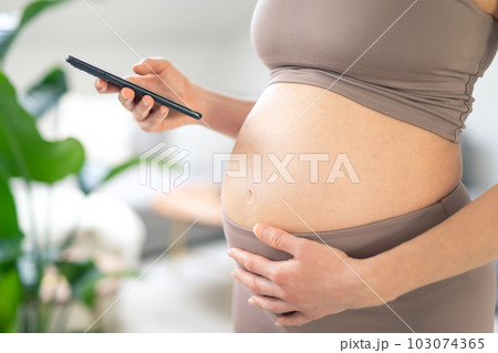 Closeup photo of pregnant female belly. Woman holding and using mobile smart phone app at home interiors. Pregnancy, technology, online shopping, preparation and expectation concept. Closeup photo of pregnant female belly. Woman holding and using mobile smart phone app at home interiors. Pregnancy, technology, online shopping, preparation and expectation concept. 103074365
