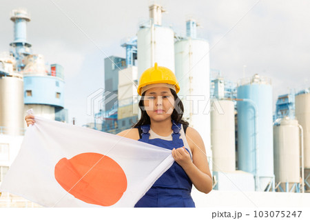 Sad young woman in helmet holding flag of Japan against background of factory 103075247