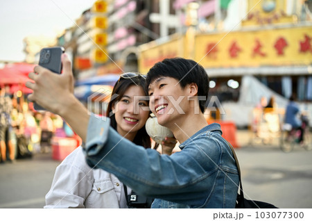 Happy young Asian tourist couple taking a photo with a smartphone in a Chinatown. 103077300