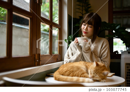 Relaxed young Asian woman sipping tea at a table with her orange cat. 103077745