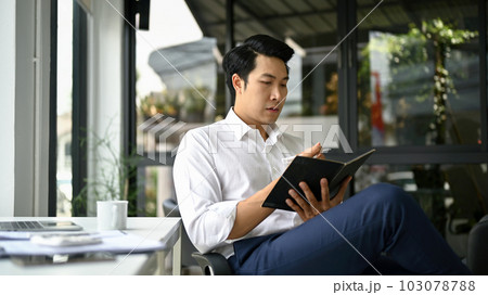 A businessman focuses on reading a book while sitting at his desk. 103078788