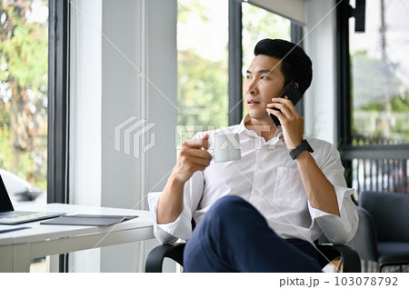 Professional Asian businessman is on the phone and sipping coffee in his office. 103078792