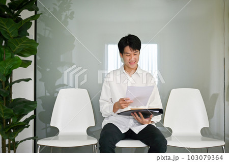 Confident Asian male candidate sits in a waiting room and rechecks his documents 103079364