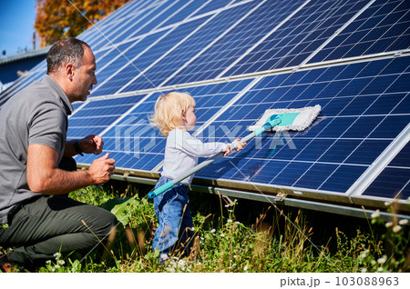 Father showing his little son the solar panels during sunny day. Cute boy helping his dad to clean solar battery. Young man teaching his small kid how to care about their source of energy. 103088963