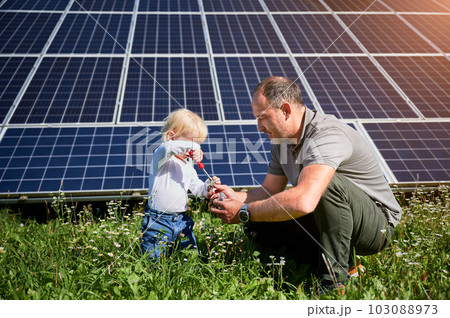 Dad showing to his little son how to use screwdriver in background of solar panels. Father teaching his child how to use tools. Son and his young dad learning tools. Dad showing to his little son how to use screwdriver in background of solar panels. Father teaching his child how to use tools. Son and his young dad learning tools. 103088973