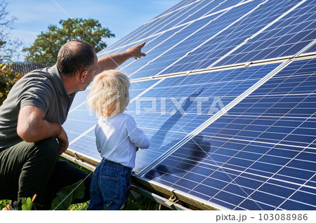 Man showing his small child the solar panels during sunny day. Father presenting to son modern energy resource. Back view of father and his kid watching on solar panels Man showing his small child the solar panels during sunny day. Father presenting to son modern energy resource. Back view of father and his kid watching on solar panels 103088986