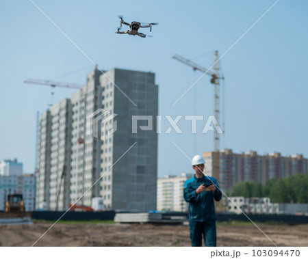 A man in a helmet and overalls controls a drone at a construction site. The builder carries out technical oversight. A man in a helmet and overalls controls a drone at a construction site. The builder carries out technical oversight. 103094470