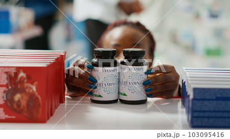 Woman organizing bottles of medicine on shelves, putting medicaments and pills on drugstore racks. Female specialist arranging vitamins and medical products. Close up. Tripod shot. 103095416
