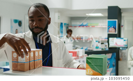 Pharmacy employee arranging stock of medicaments on drugstore shelves, putting boxes of pills and vitamins to help customers. Young man working with pharmaceutical products, retail store. 103095450