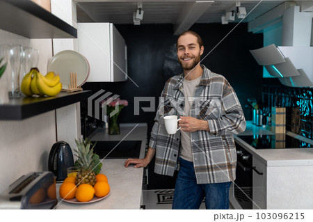 Man with beard holding cup with coffee, standing by kitchen counter with modern light interior. 103096215