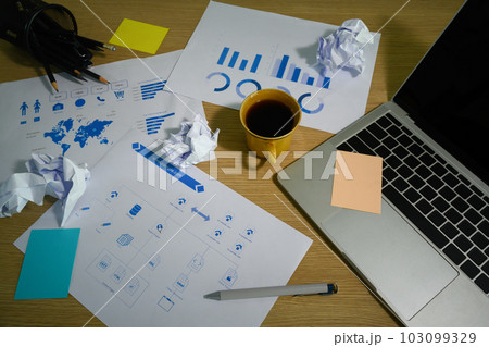 Cluttered office desk with crumpled papers, laptop, coffee cup and sticky notes 103099329