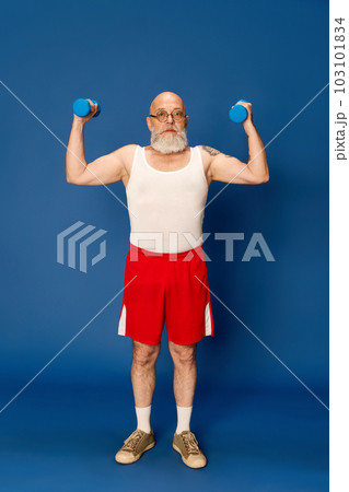 Portrait of mature, bearded man in sportswear training with dumbbells against blue studio background 103101834