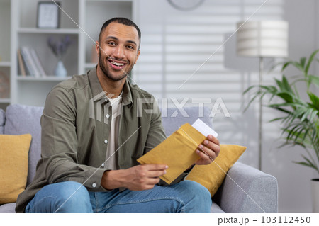 Portrait of a young Muslim man smiling looking at the camera and holding a letter, documents in an envelope delivered to his home. 103112450
