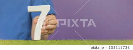Number 7 in hand. A hand holds a white number seven on green grass and a purple background, seven percent per annum. World Health Day. April 7th. Holiday concept. Number 7 in hand. A hand holds a white number seven on green grass and a purple background, seven percent per annum. World Health Day. April 7th. Holiday concept. 103115738