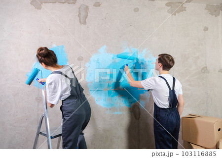 House decoration DIY renovation concept. Happy couple in empty room of new home painting wall together. Husband and wife holding paint roller painting wall with blue color paint in new house House decoration DIY renovation concept. Happy couple in empty room of new home painting wall together. Husband and wife holding paint roller painting wall with blue color paint in new house 103116885