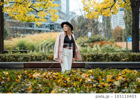Fashion portrait of stylish young woman in hat walking in the autumn city. Happy gen z woman walking in the city with yellow autumn foliage 103118403