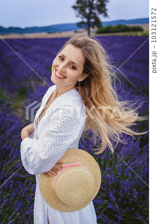 In a lavender field, a beautiful woman is standing with a hat in her hand in the middle of the flower 103121572