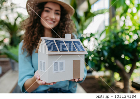 Young woman in botanical garden holding model of house with solar panels, concept of green energy. Young woman in botanical garden holding model of house with solar panels, concept of green energy. 103124384