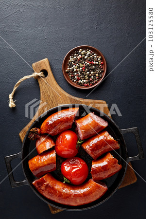 Fried pork sausage cut into pieces in a cast iron pan with tomatoes and peppercorns on a wooden cutting board 103125580