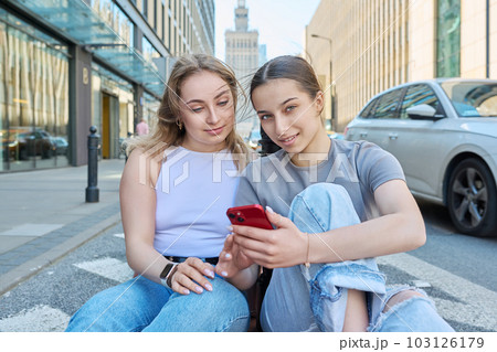 Two smiling female students with smartphone, modern urban style, road, buildings background 103126179