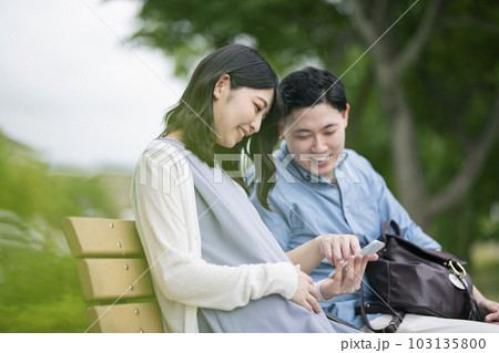 公園でスマートフォンを見る若い夫婦 公園でスマートフォンを見る若い夫婦 103135800