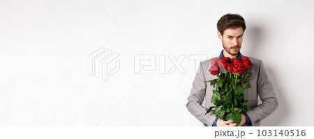 Handsome macho man going on date in suit, holding bouquet of red roses and smiling at camera, making valentines day surprise gift, white background 103140516