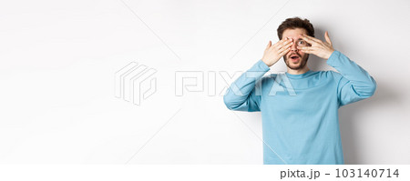 Amazed smiling man covering eyes with hands but peeking through fingers at something awesome, standing over white background Amazed smiling man covering eyes with hands but peeking through fingers at something awesome, standing over white background 103140714