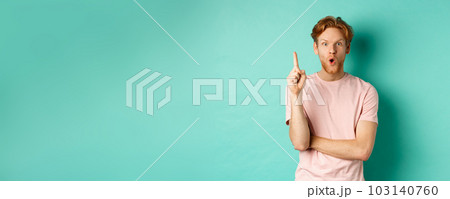 Pensive redhead man in t-shirt raising index finger, gasping as pitching at idea, saying suggestion, standing over turquoise background Pensive redhead man in t-shirt raising index finger, gasping as pitching at idea, saying suggestion, standing over turquoise background 103140760