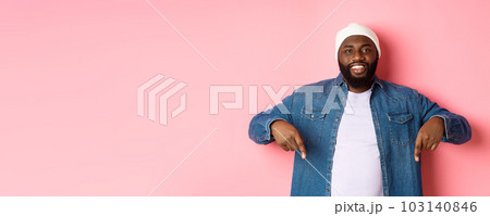 Handsome hipster african-american man pointing fingers down, smiling and showing promo offer, standing over pink background 103140846