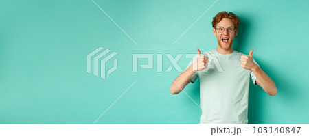Excited young man with red hair, wearing glasses, showing thumbs-up and agree or praise something, smiling amazed and saying yes, standing over turquoise background 103140847