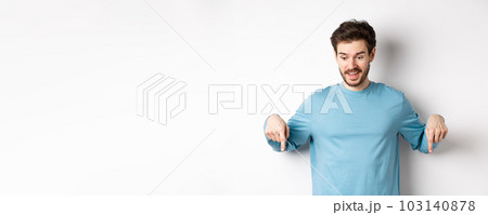 Excited and happy young man looking and pointing down at awesome deal, checking out promotion offer, standing on white background Excited and happy young man looking and pointing down at awesome deal, checking out promotion offer, standing on white background 103140878