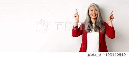 Happy mature woman in red blazer and makeup, smiling and showing advertisement on top, pointing fingers up at logo, white background Happy mature woman in red blazer and makeup, smiling and showing advertisement on top, pointing fingers up at logo, white background 103140910