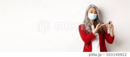 Covid, pandemic and business concept. Businesswoman in red blazer and face mask using hand sanitizer to clean and disinfect, standing over white background 103140922