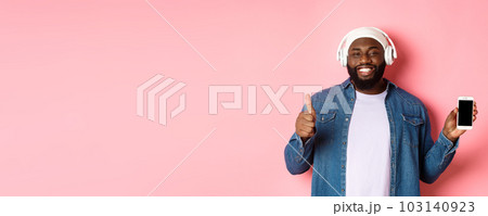Happy hipster guy listening music on headphones and showing mobile screen, smiling satisfied, showing thumb-up in approval, like playlist, standing over pink background Happy hipster guy listening music on headphones and showing mobile screen, smiling satisfied, showing thumb-up in approval, like playlist, standing over pink background 103140923