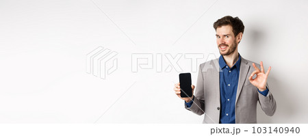 E-commerce and online shopping concept. Smiling businessman show okay sign and empty smartphone screen, demonstrate his account, standing on white background 103140940