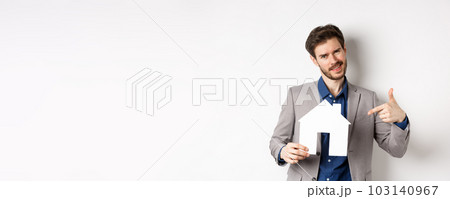 Real estate and insurance concept. Salesman in grey suit showing paper house cutout, selling property, smiling at camera, white background 103140967