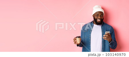 Image of stylish Black man hipster drinking takeaway coffee, reading message on phone and smiling, standing over pink background Image of stylish Black man hipster drinking takeaway coffee, reading message on phone and smiling, standing over pink background 103140970
