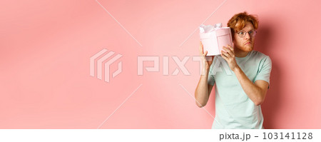 Love and holidays concept. Intrigued redhead guy press ear to box and shaking gift, guessing what inside, standing over pink background 103141128