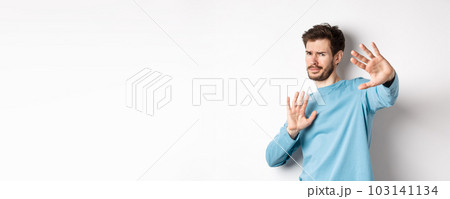 Stay away from me. Reluctant young man step back with hands stretch out in defensive gesture, protecting himself, standing over white background 103141134