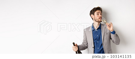 Celebration and holidays concept. Handsome bearded man in suit and birthday hat holding bottle, drinking glass of champagne, standing on white background 103141135