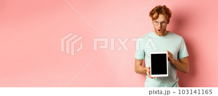 Impressed redhead man in glasses, showing blank digital tablet screen and looking in awe at display, saying wow, standing in t-shirt against pink background 103141165
