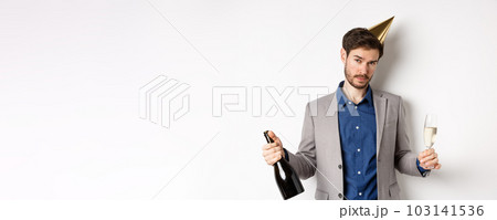 Party guy standing in birthday hat and celebrating, holding champagne bottle and glass, looking drunk, wearing grey suit 103141536