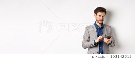 Handsome bearded male model in suit using mobile phone, smiling pleased at camera, white background 103142613
