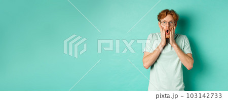 Shocked redhead guy in glasses checking out awesome promotion offer, gasping amazed and holding hands on face, staring at camera, mint background 103142733