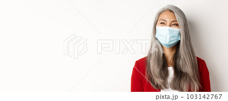 Covid, pandemic and business concept. Close up of happy asian businesswoman with grey hair, wearing medical mask and smiling, looking left with cheerful face, white background 103142767