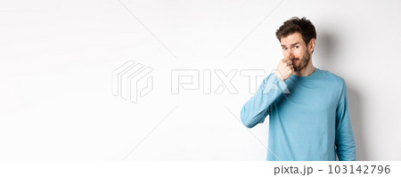 Disgusted young man in sweatshirt shut his nose, looking at something with bad smell, standing over white background Disgusted young man in sweatshirt shut his nose, looking at something with bad smell, standing over white background 103142796